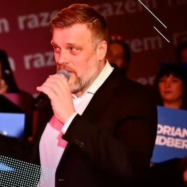 Adrian Zandberg przemawiający na wiecu politycznym lub konferencji prasowej, z dynamiczną postawą, podkreślającą jego zaangażowanie.