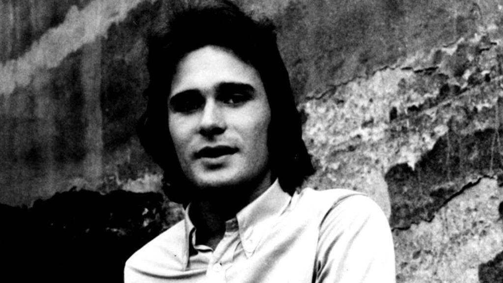 Jerzy Grunwald w energetycznym ujęciu podczas występu na scenie w latach 70., grający na gitarze lub śpiewający do mikrofonu, z nastrojowym oświetleniem scenicznym.