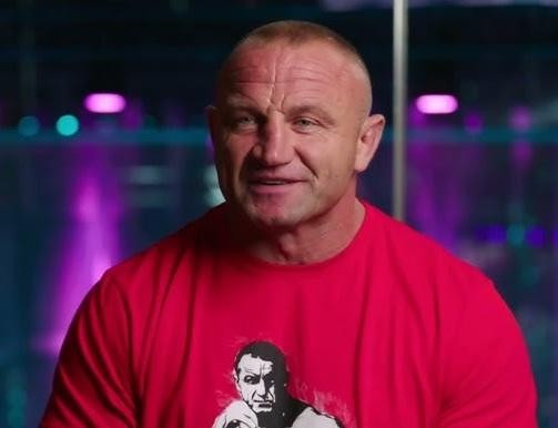 Mariusz Pudzianowski w postawie bojowej MMA, z zaciętym wyrazem twarzy, na tle rozmytej widowni oktagonu.